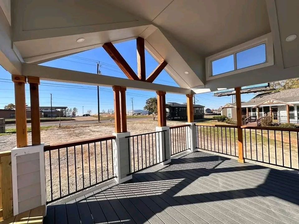 Covered wooden deck with tall beams, black railings, and slanted roof. Overlooks an open yard under a clear blue sky, evoking a peaceful vibe.