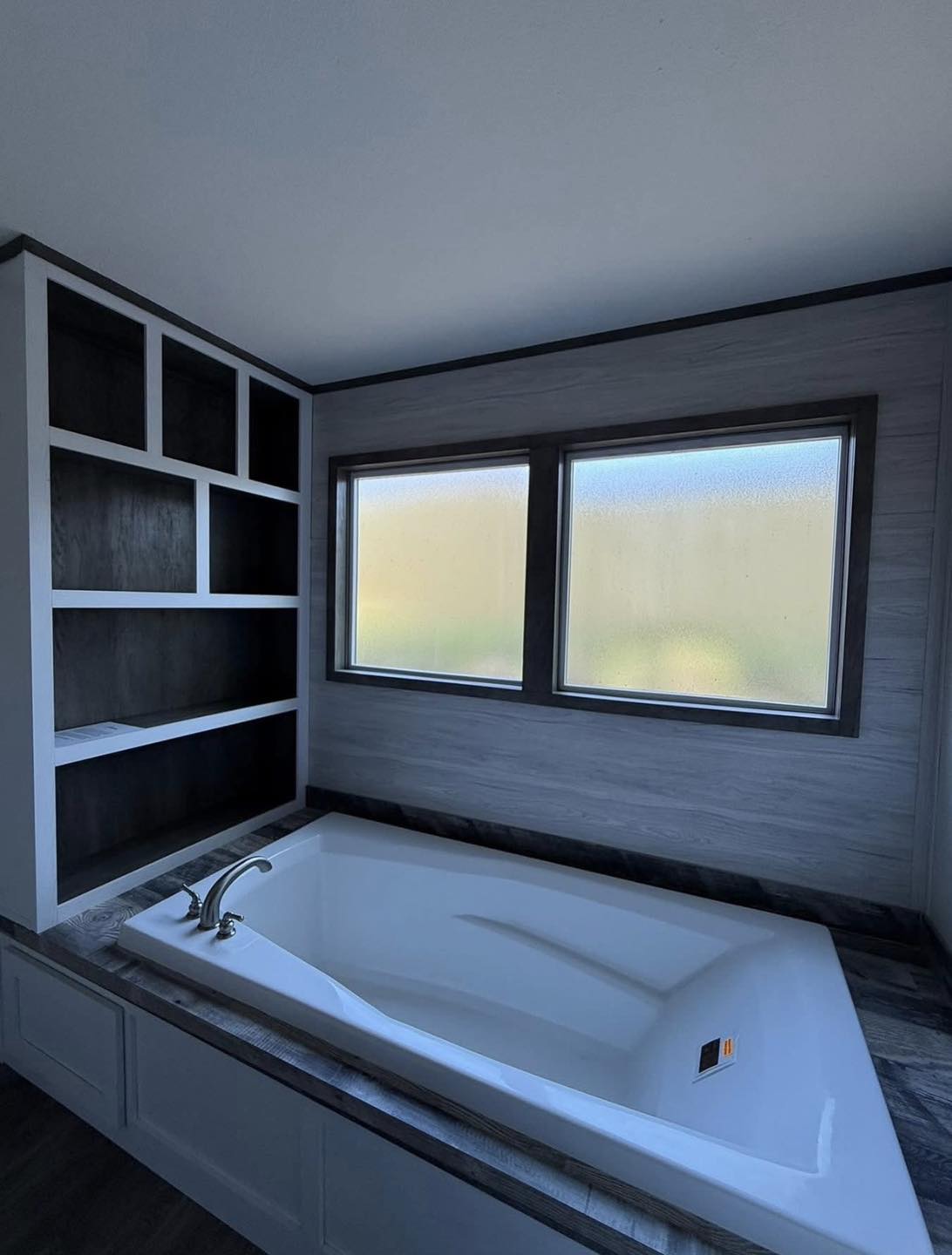 Modern bathroom with a large white bathtub set in dark wood. Two frosted windows above, and sleek shelving to the left. Calm and minimalist ambiance.