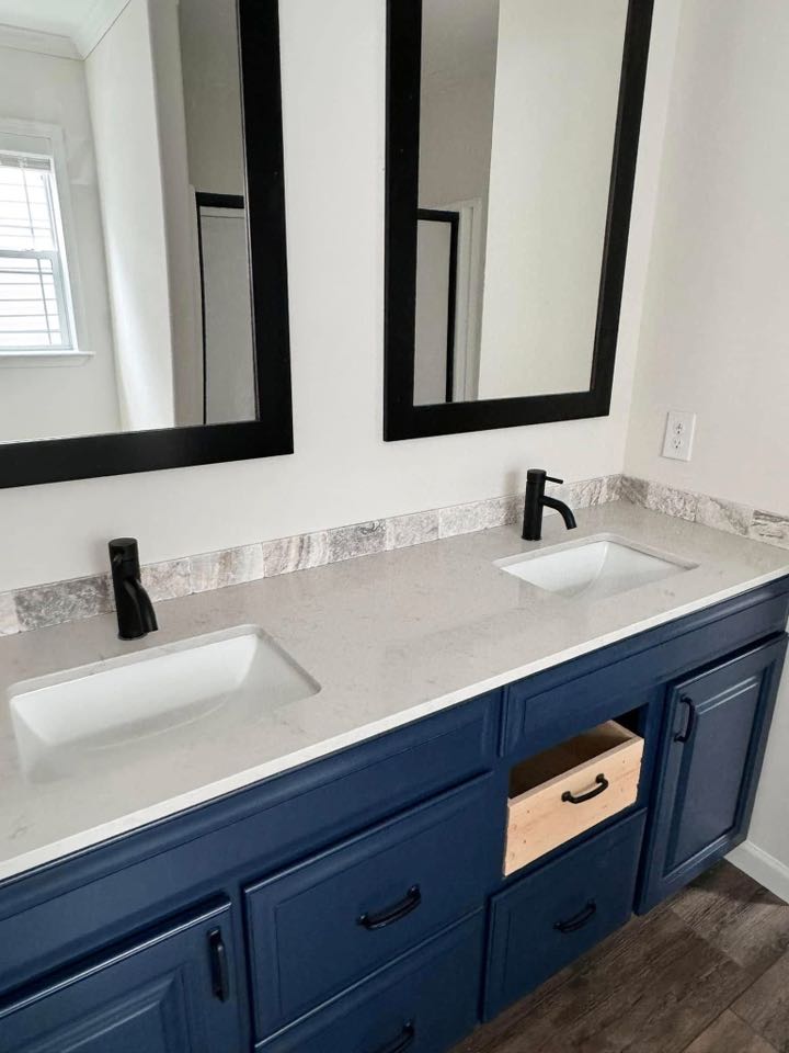 Modern bathroom with a double sink vanity. Features a white countertop, black faucets, and blue cabinets, with two large mirrors above. One drawer is open.