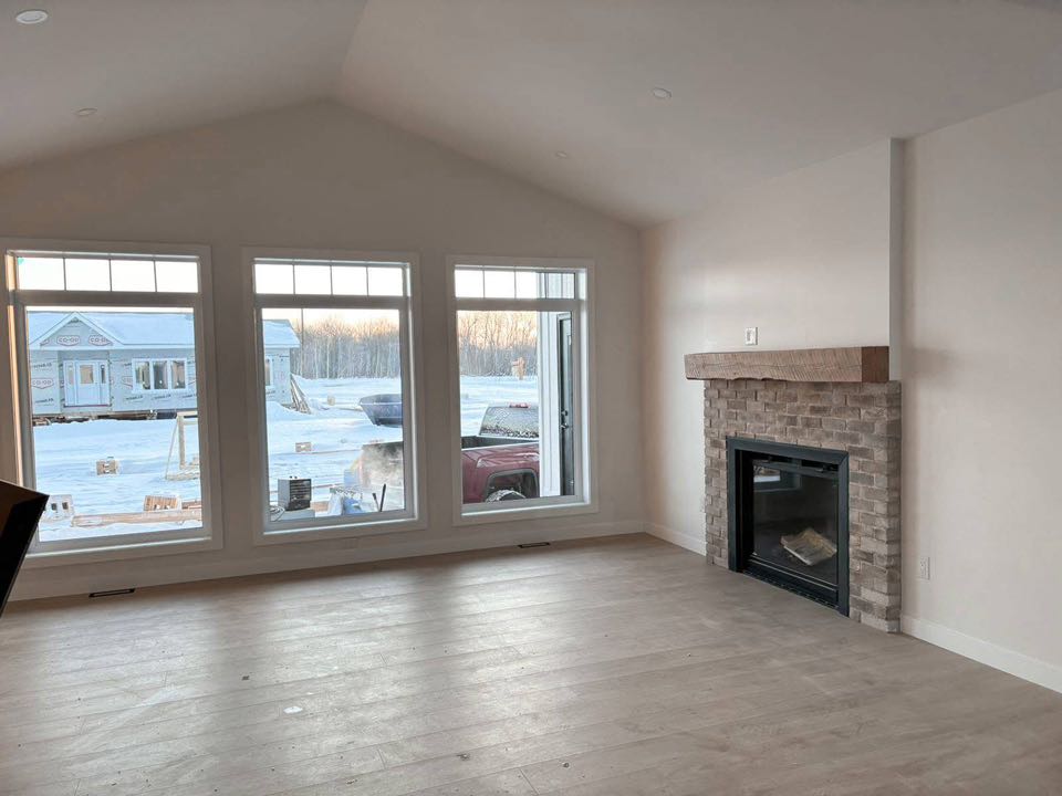 Spacious room with large windows showing a snowy yard, sunlight warming the space. A brick fireplace adds a cozy touch, enhancing the serene atmosphere.