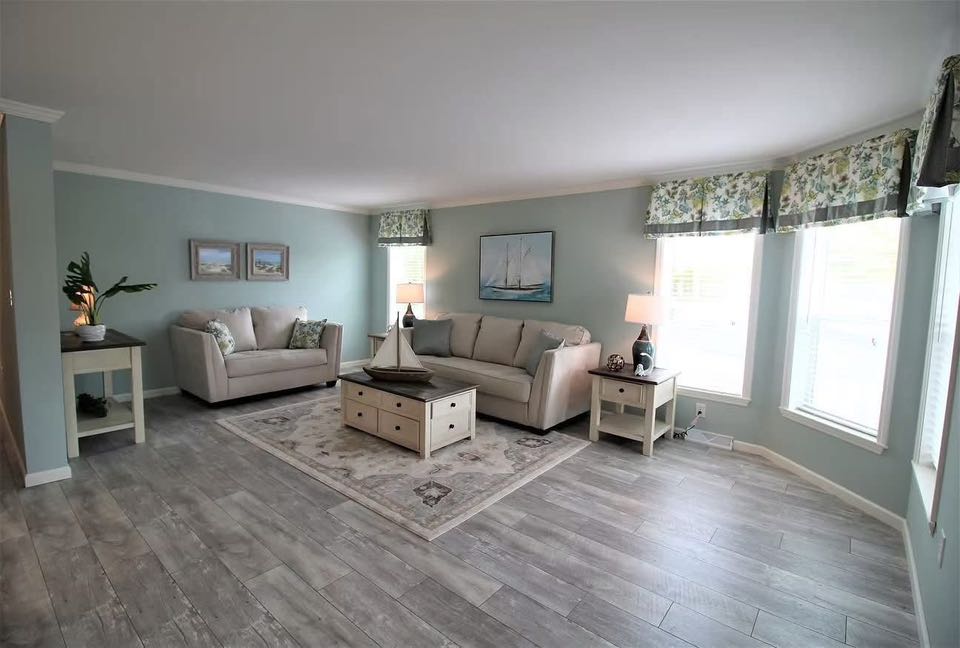 Spacious living room with light gray wooden floors, soft blue walls, two beige sofas, nautical decor, floral curtains, and large windows. Bright and serene.