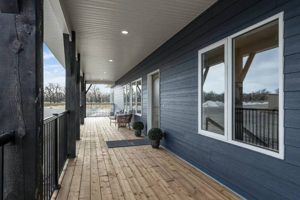 Spacious wooden porch with blue siding, two black railings, and a wicker chair set. Overhead lights and potted plants add a welcoming touch.
