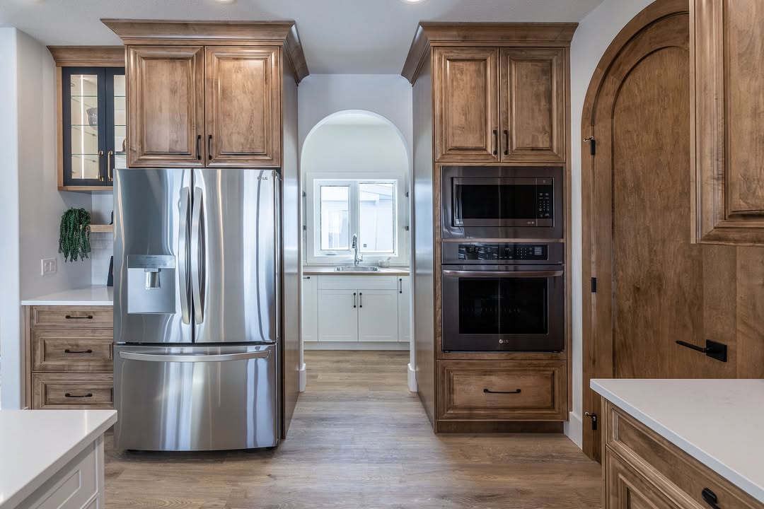 Modern kitchen with wooden cabinets, a stainless steel refrigerator, and built-in double ovens. An arched doorway leads to a bright sink area.