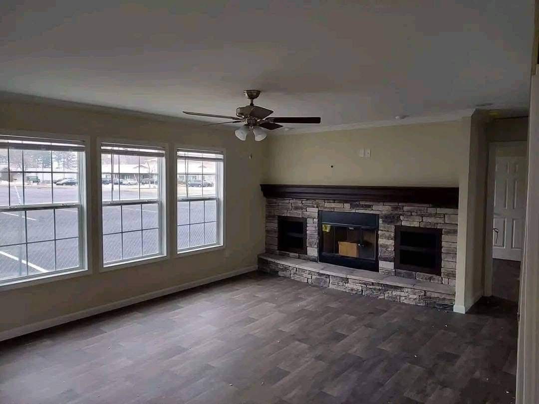 Bright living room with three large windows, a stone fireplace, and a ceiling fan. Hardwood floors add warmth to the spacious, inviting area.