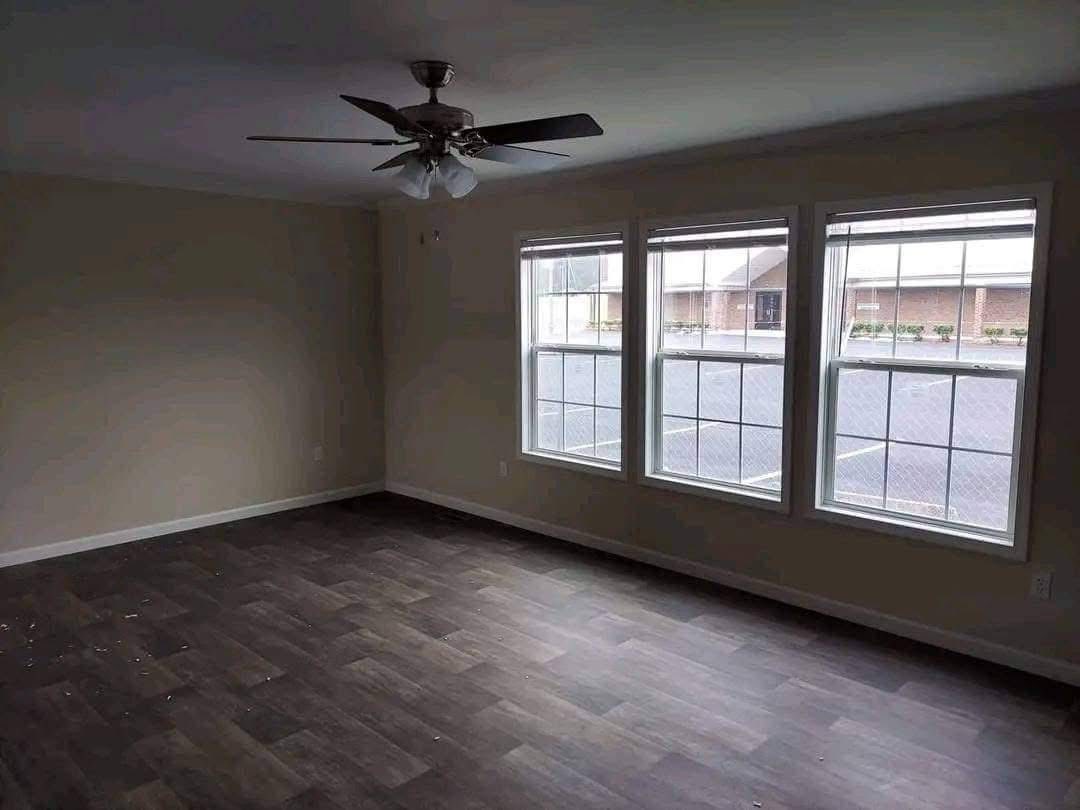 Empty room with light beige walls and dark wood flooring. Three large windows brighten the space, and a ceiling fan is centrally mounted. The atmosphere is calm and inviting.