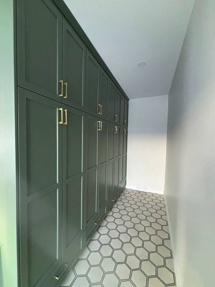 Narrow hallway with dark green cabinets and gold handles on the left. The floor features hexagonal tiles, and the right wall is plain white.
