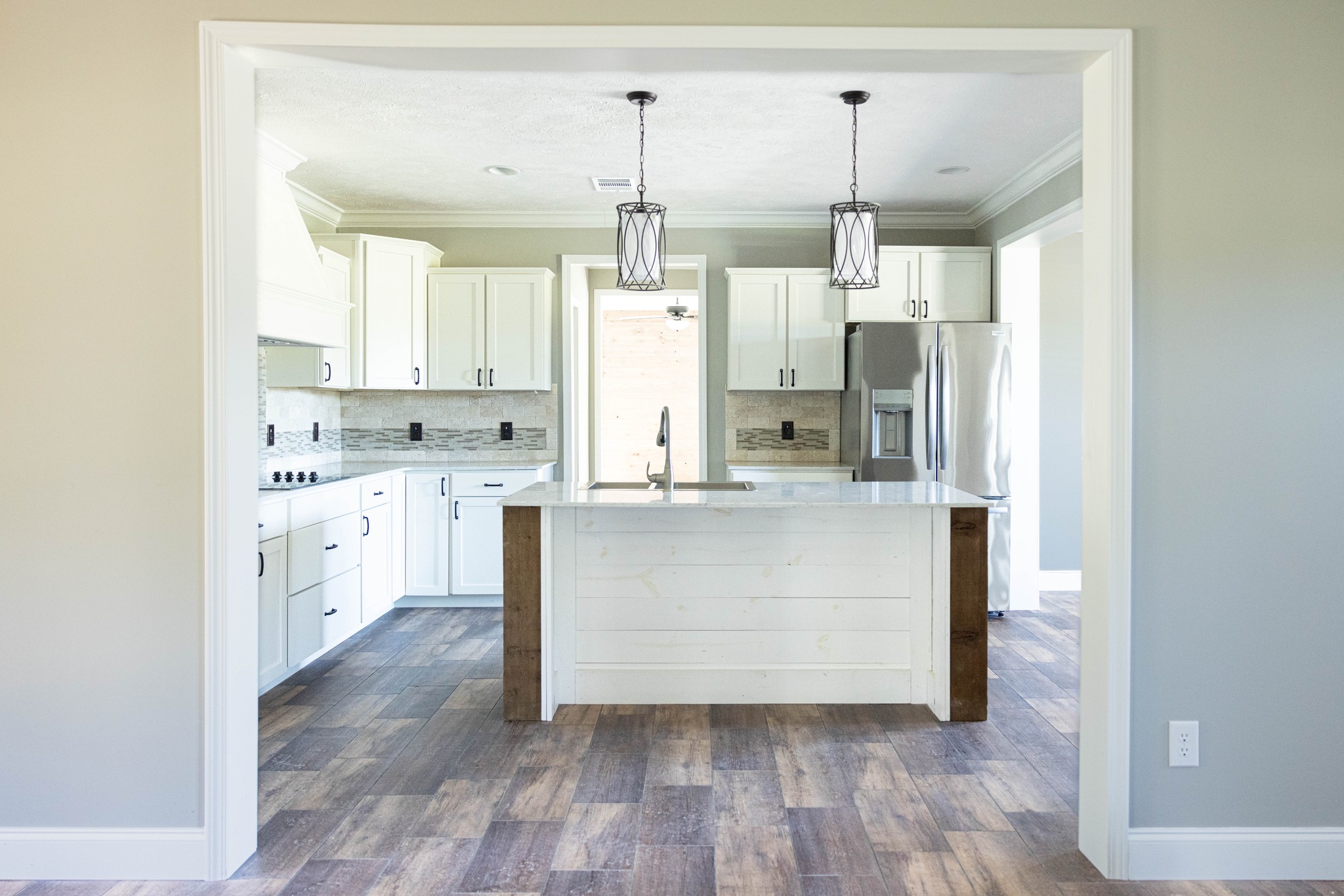 Modern kitchen with white cabinets, marble-topped island, and wood accents. Two pendant lights hang above, with stainless steel appliances. Bright and spacious.