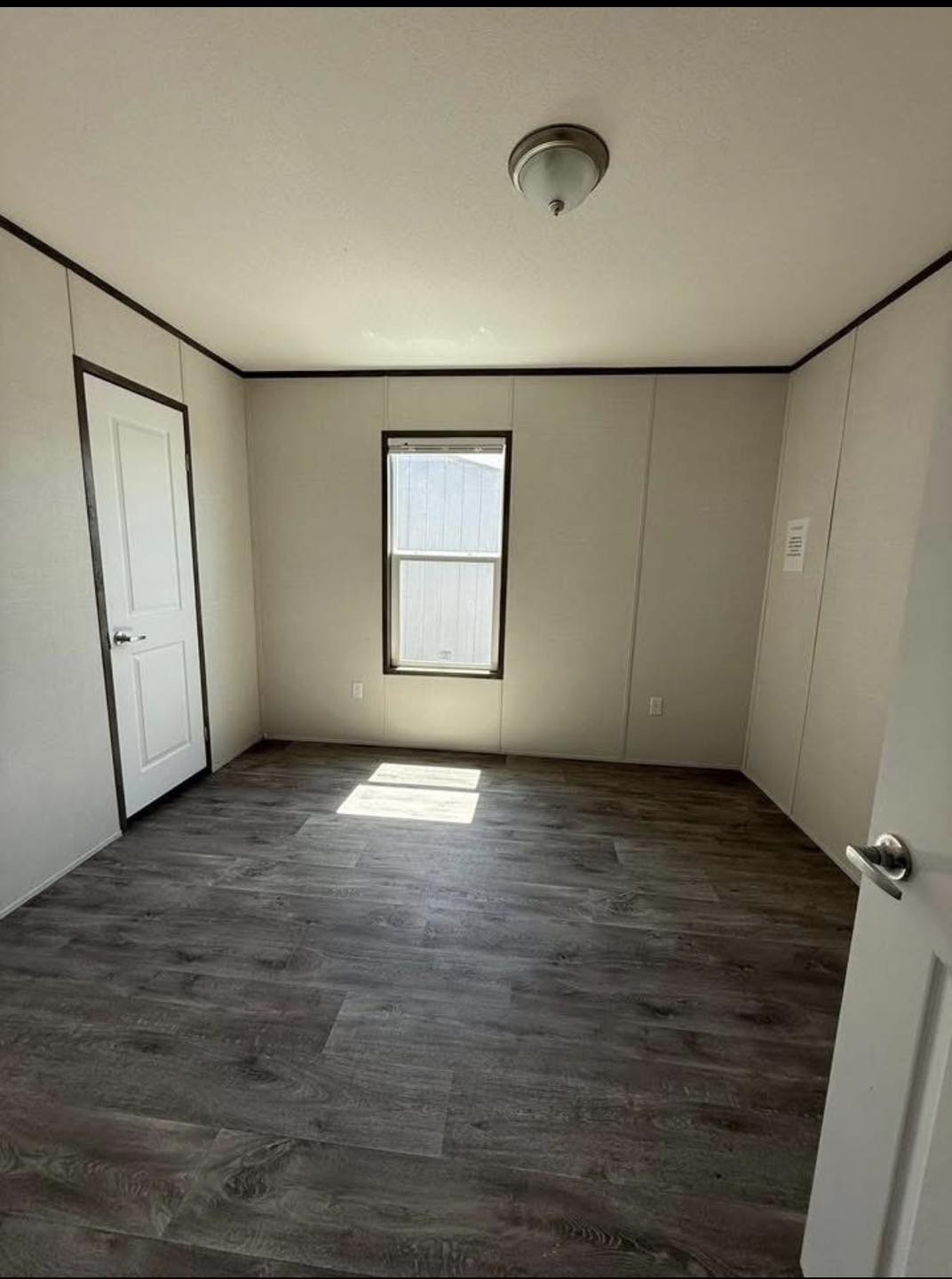Empty room with gray wooden flooring, beige walls, and a single window letting in daylight. Two closed doors and a ceiling light fixture are visible.