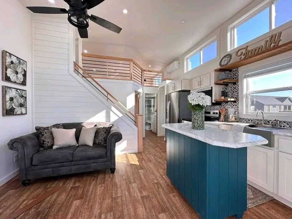 Stylish, cozy interior of a tiny home with a modern kitchen. Features a blue island, gray sofa, white walls, and a staircase leading to a loft. Warm and inviting.