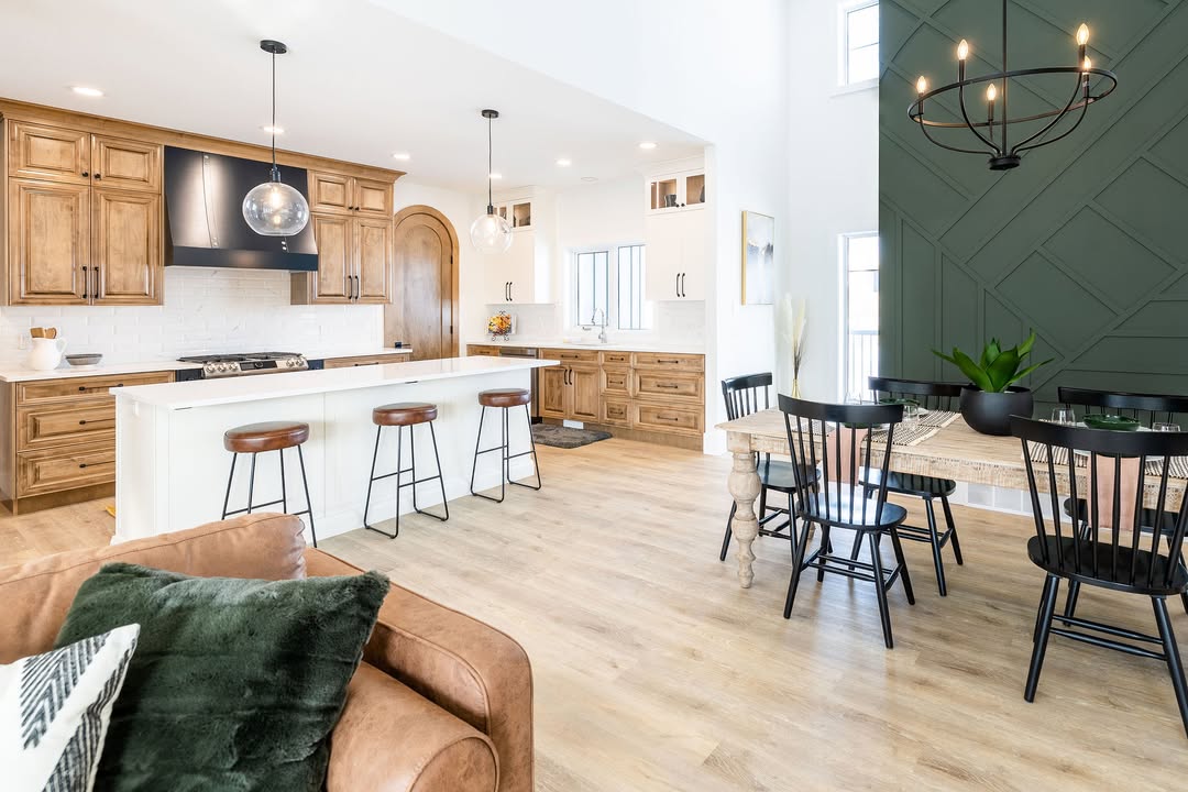 Modern kitchen with wood cabinets, white island, and three barstools. Dining area features a dark green accent wall and round chandelier. Cozy, open space.