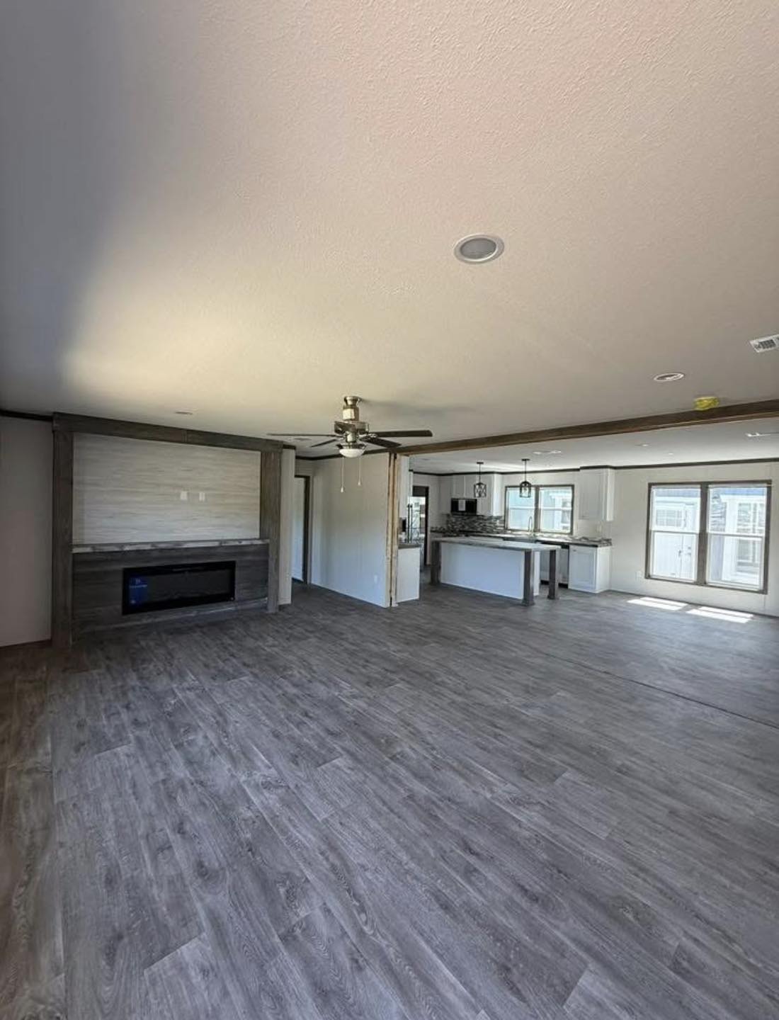 Spacious, sunlit open-plan room with grey wood floors. Features a modern fireplace, ceiling fan, and adjoining kitchen with island, two large windows.