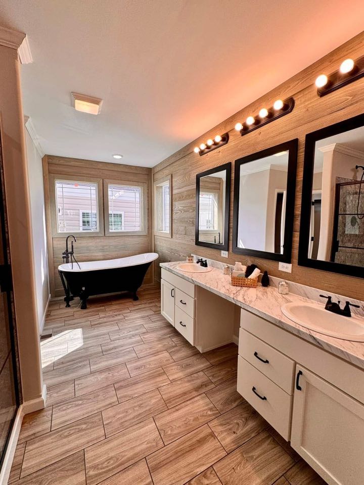 Modern bathroom with wood flooring and walls, featuring a black freestanding tub by a window. Dual sinks with black-framed mirrors are brightly lit.