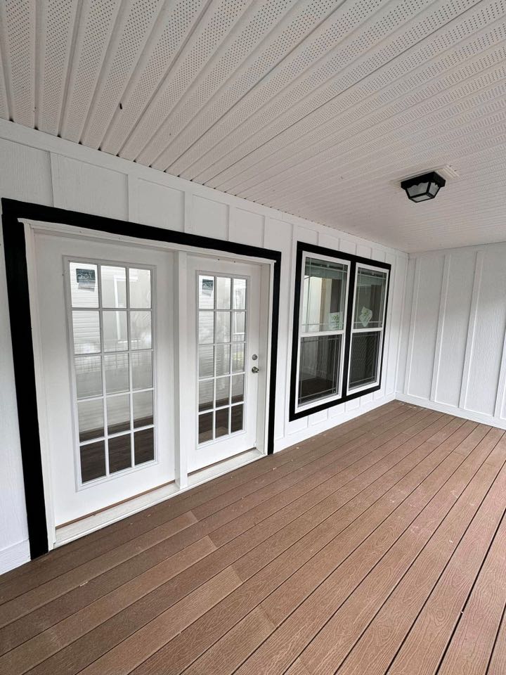 Covered porch with white paneled walls and ceiling, featuring glass French doors and large windows with black trim. Wood-grain flooring adds warmth.