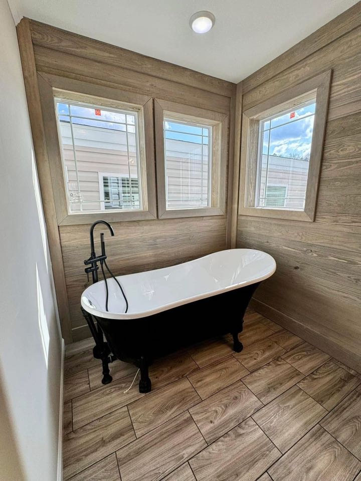 A cozy bathroom with a black clawfoot bathtub, rustic wood paneling, and three large windows. The space feels serene and inviting, with natural light.