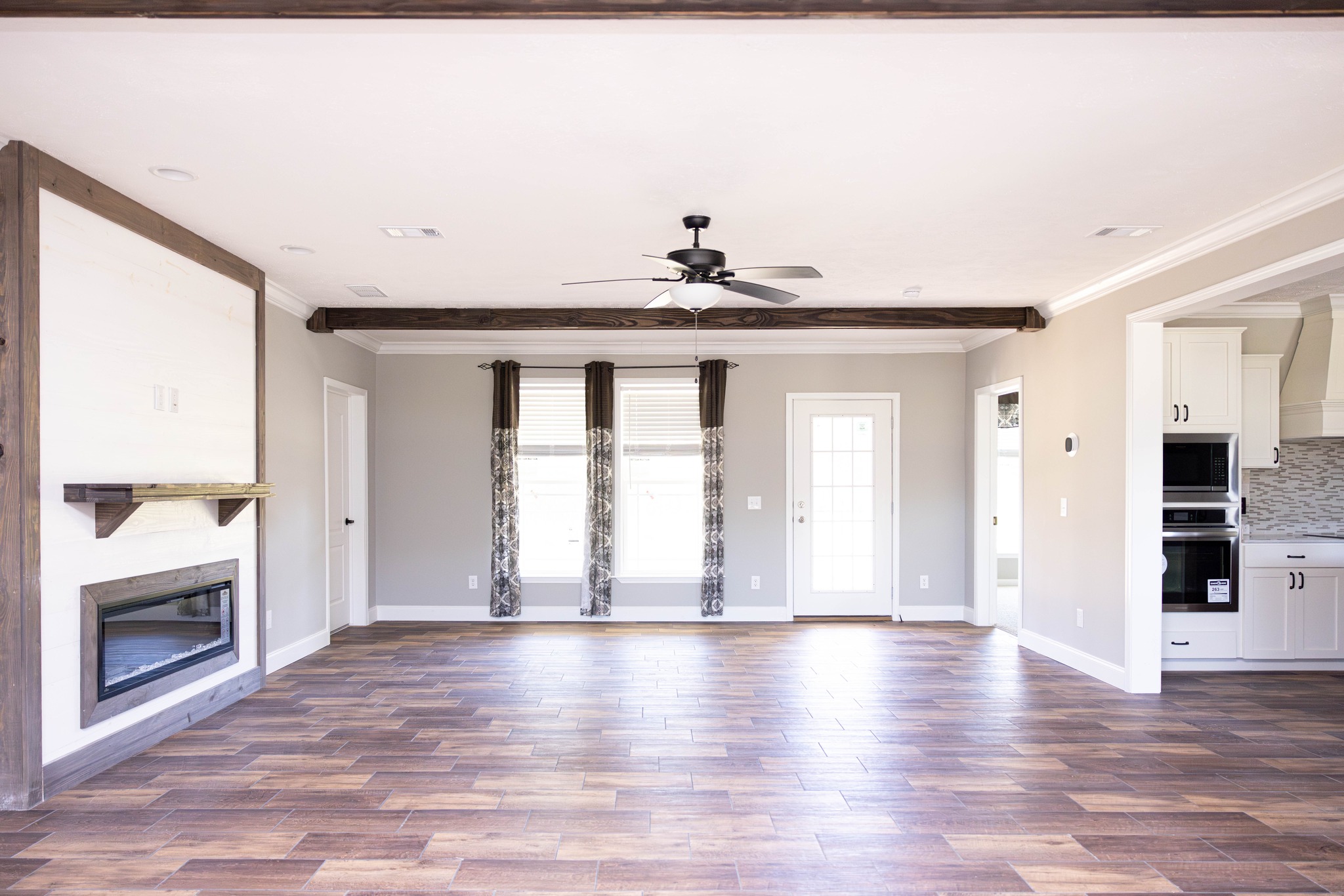 Spacious modern living room with wood flooring, large windows with patterned curtains, a ceiling fan, built-in fireplace, and view into a white kitchen.