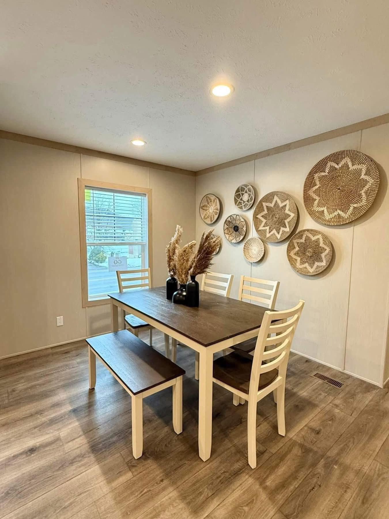 A dining room with a wooden table and white chairs, featuring a pampas grass centerpiece. Decorative woven baskets adorn the beige wall, creating a cozy ambiance.