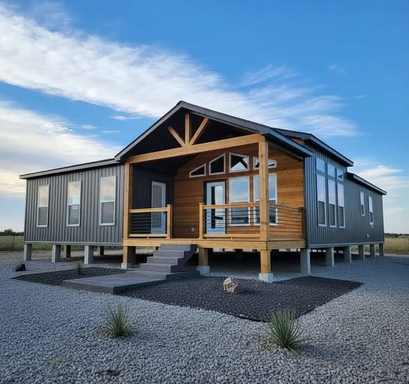 Modern home on stilts with a wooden porch and metal siding sits in a gravel landscape. The sky is clear with a few clouds, creating a serene atmosphere.