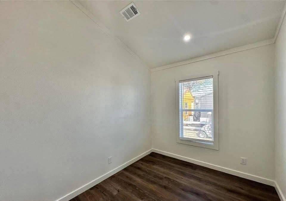 Small, empty room with white walls and a sloped ceiling. A single window on the right shows a glimpse of a yellow house outside. Dark wooden floor.