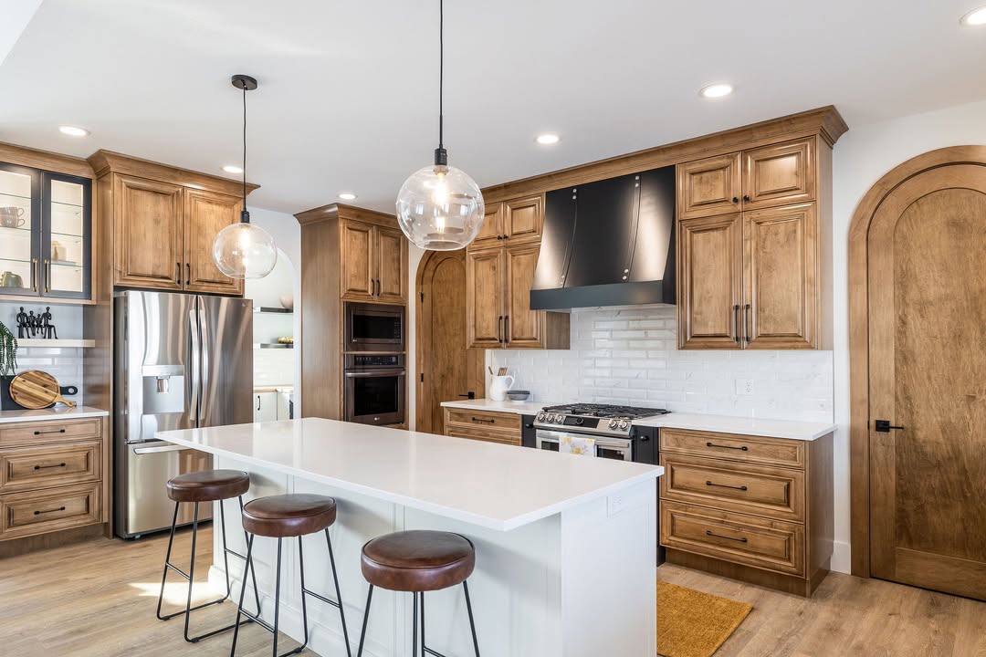 Modern kitchen with wooden cabinets, stainless steel appliances, and a white island with three brown stools. Glass pendant lights hang above, creating a warm, inviting tone.