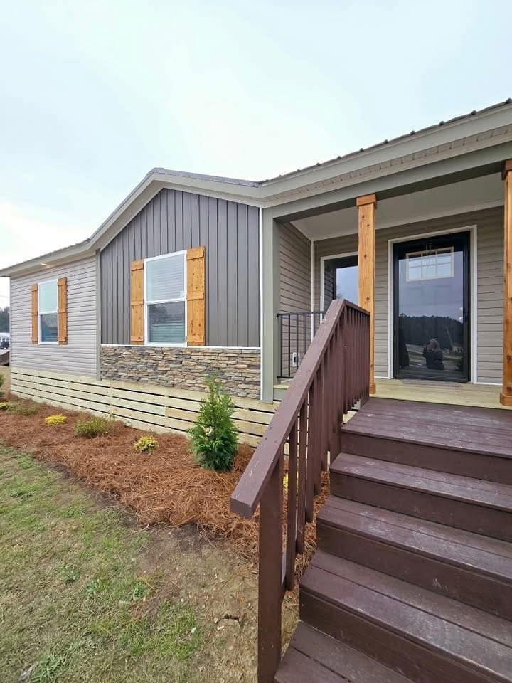 Alt text: "Modern single-story home with gray siding, stone accent, and wooden shutters. Brown steps lead to the porch, surrounded by fresh landscaping."
