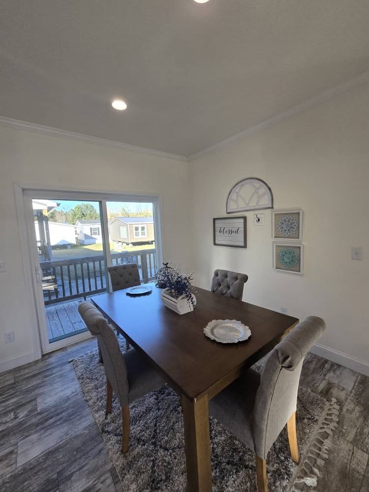 A cozy dining room with a wooden table, four tufted gray chairs, and a floral centerpiece. Bright light enters through a sliding glass door, enhancing the inviting atmosphere. Wall art includes a "Blessed" sign and decorative frames.