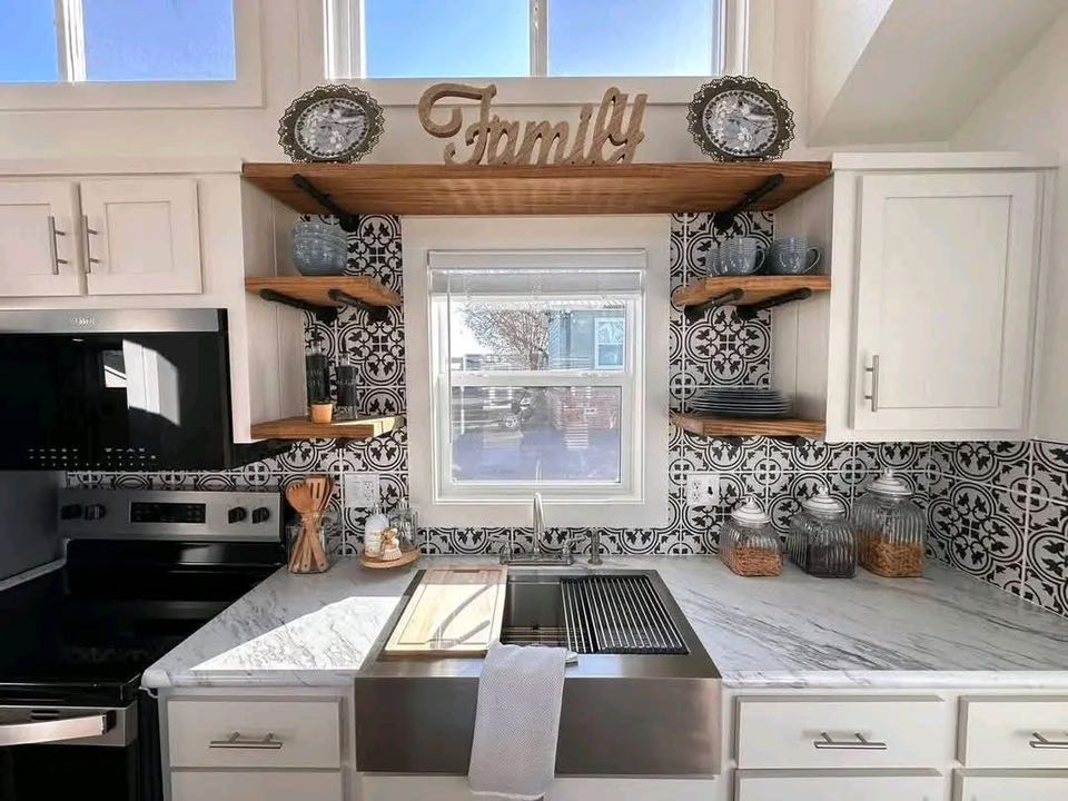 Bright kitchen with a farmhouse sink, black and white patterned backsplash, and wooden shelves displaying dishes. "Family" sign adds warmth.