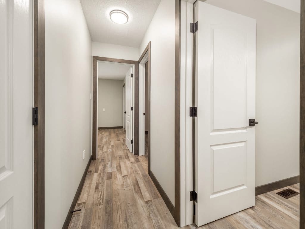 A bright hallway with wooden floors and light gray walls. Several white doors with dark frames are open, leading to rooms, evoking a welcoming feel.