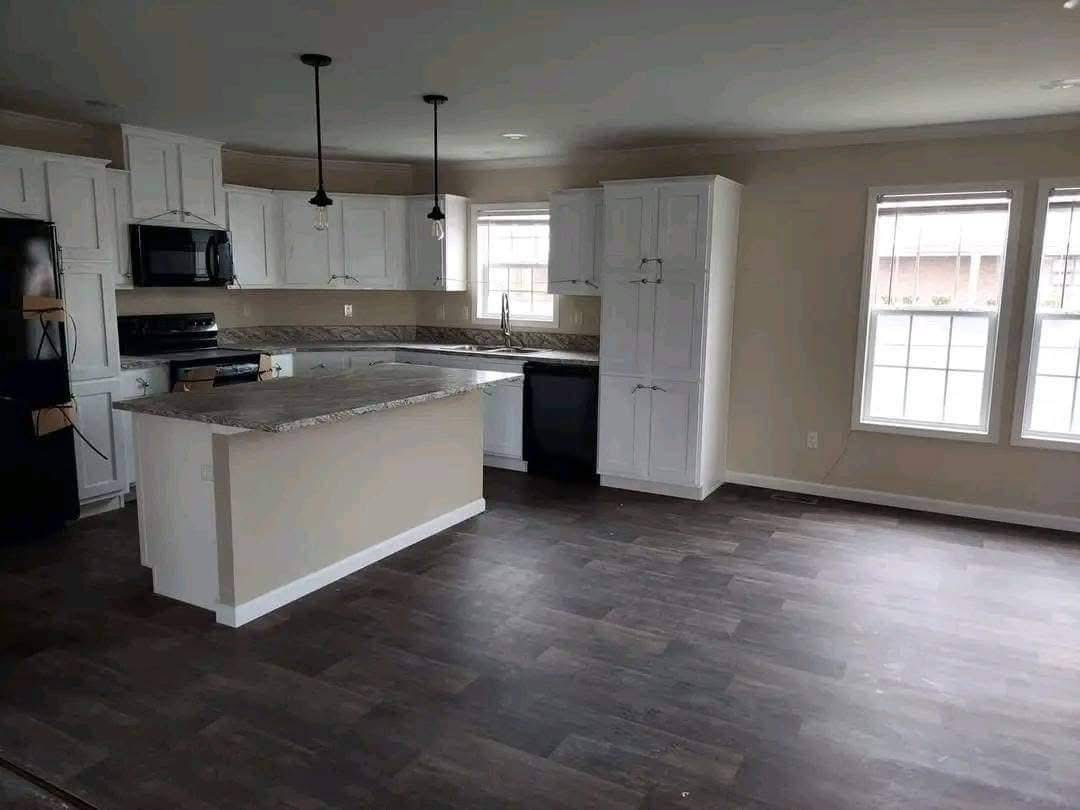 Spacious kitchen with white cabinets, dark wood floor, and a large island. Two pendant lights hang above. Sunlight streams through windows, creating a cozy feel.