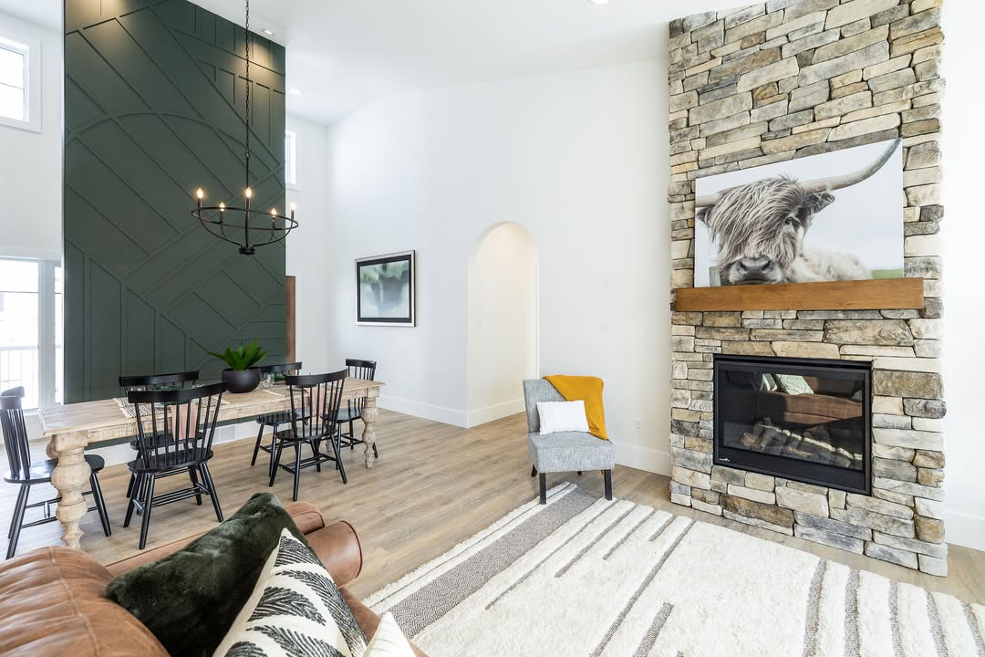 Modern living room with stone fireplace, featuring a cow artwork. Wooden dining table under chandelier, green accent wall, and cozy seating area. Warm and inviting ambiance.