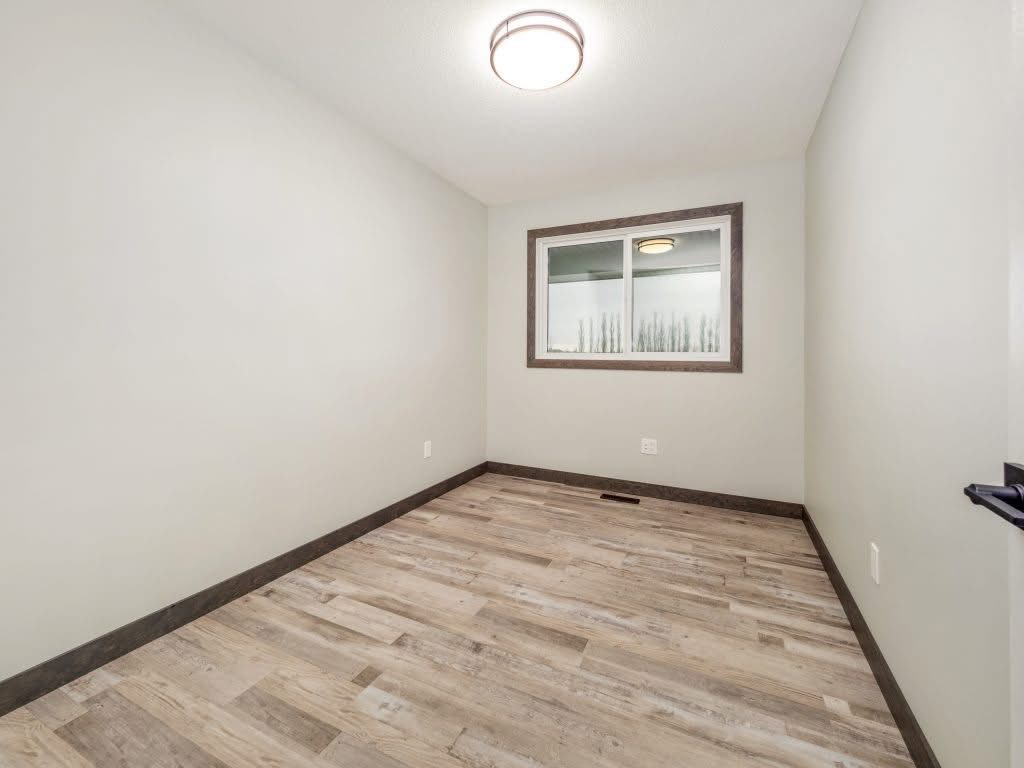 Empty room with light gray walls, a small window with a dark trim, and wood-look laminate flooring. Ceiling light casts a soft glow, creating a calm atmosphere.