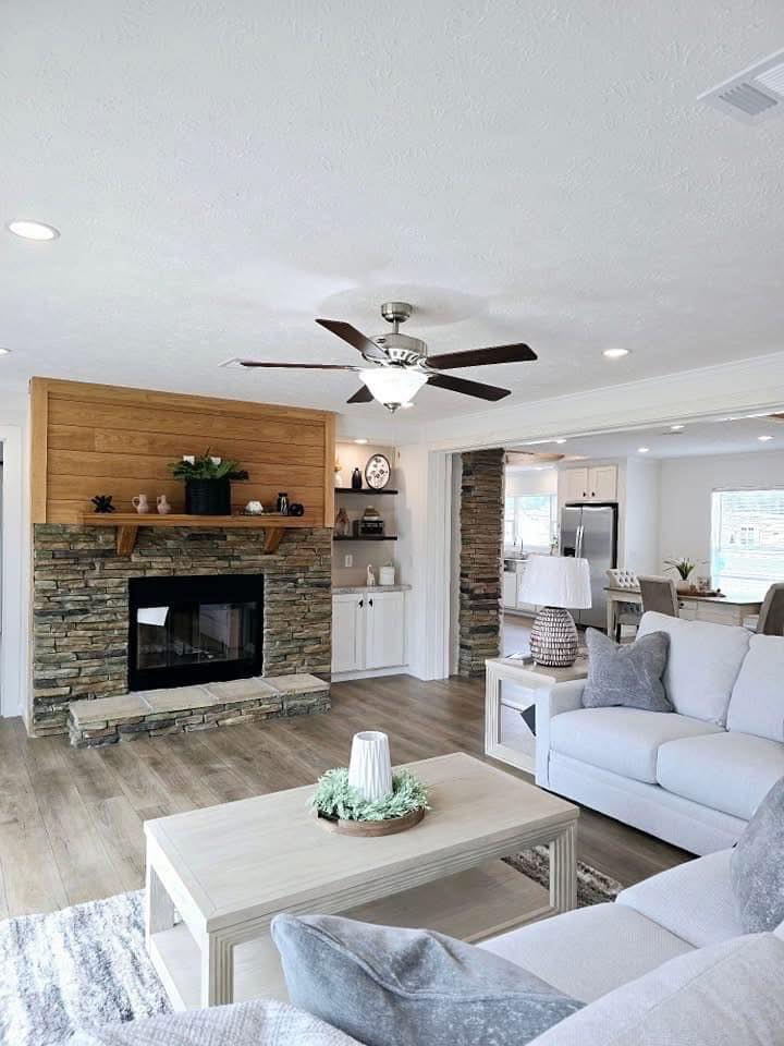 Spacious living room with light gray sofas, a stone fireplace, and a light wood coffee table. Ceiling fan overhead, bright ambiance, open kitchen view.