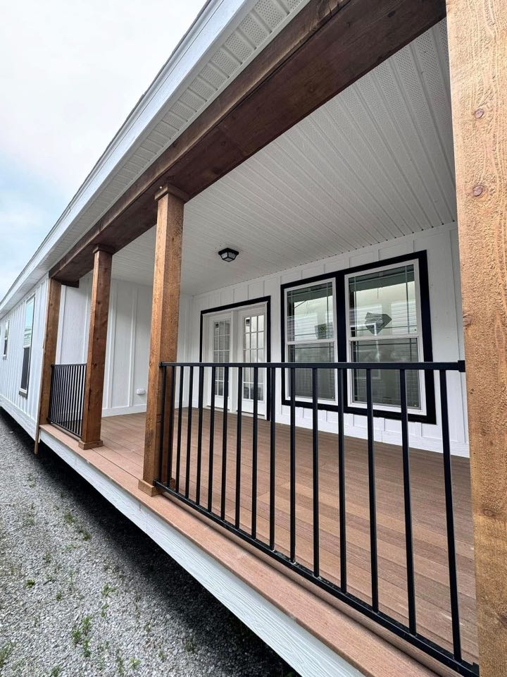 Wooden porch with natural wood beams and black railing on a white house. The ceiling is white, and windows with dark trim face the porch.