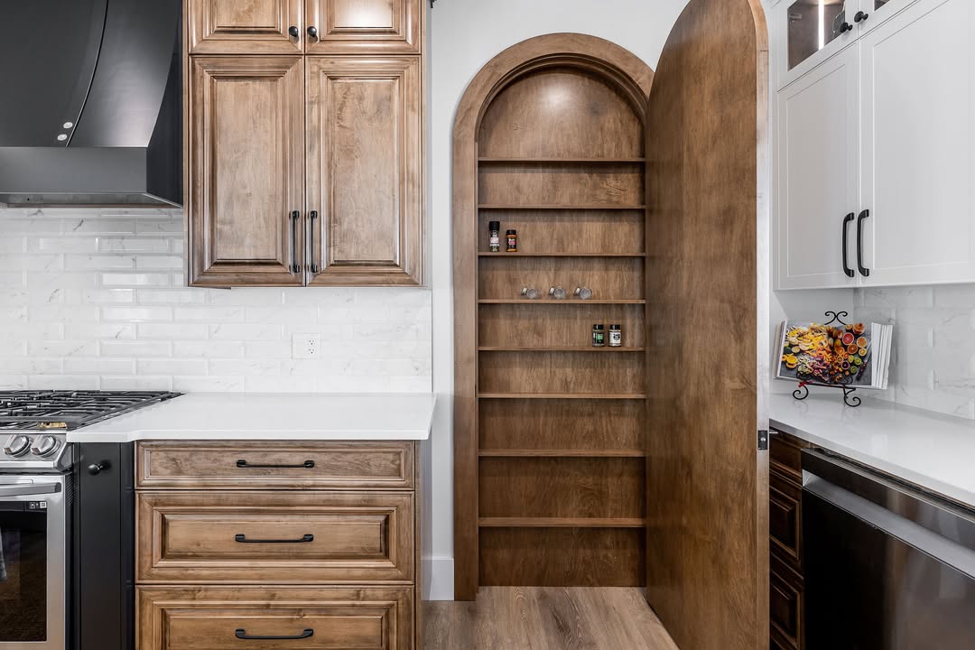 Modern kitchen with light wood cabinetry and sleek, white countertops. A secret door opens to reveal a pantry with arched shelves, creating intrigue.