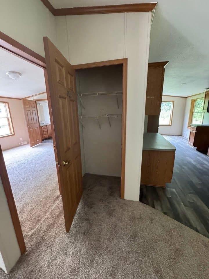 Open closet with wooden door and empty shelves on carpeted floor. Nearby are carpeted and wood-floored rooms with windows, creating an empty, spacious feel.