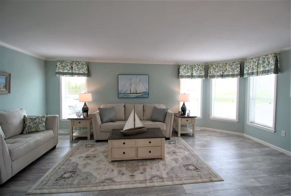 Cozy living room with light teal walls, floral curtains, and wooden flooring. A sailboat model sits on the coffee table. Two beige sofas face each other, flanking a central painting of a sailboat, creating a serene nautical theme.
