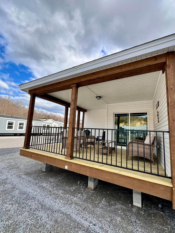 Covered wooden porch with wicker chairs and a railing, attached to a house. The sky is partly cloudy, and nearby homes are visible, suggesting a peaceful neighborhood.