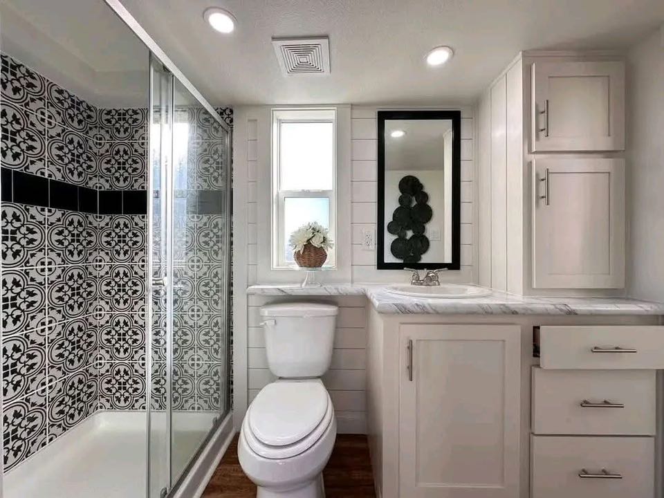 Compact modern bathroom with patterned black-and-white shower tiles, white fixtures, and wood floor. A potted plant sits on the windowsill.