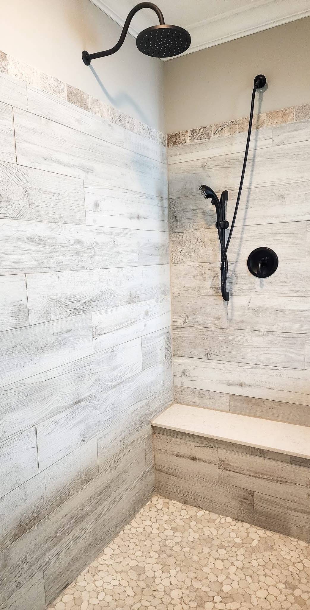 A modern shower with wood-textured tiles, pebble stone floor, a black rainfall showerhead, and a hand-held shower handle next to a built-in bench.