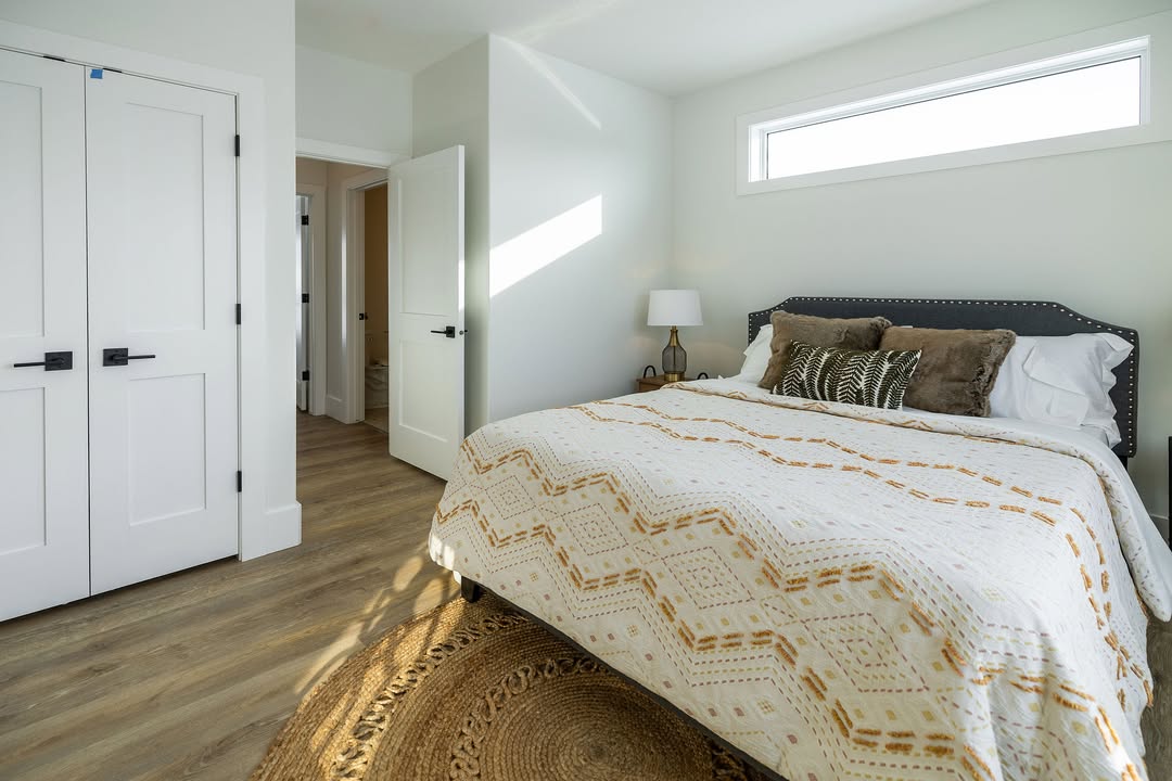 Bright bedroom with a sunlit bed covered in a geometric-patterned blanket, white walls, hardwood floor, and a small lamp on a side table. Cozy and serene.