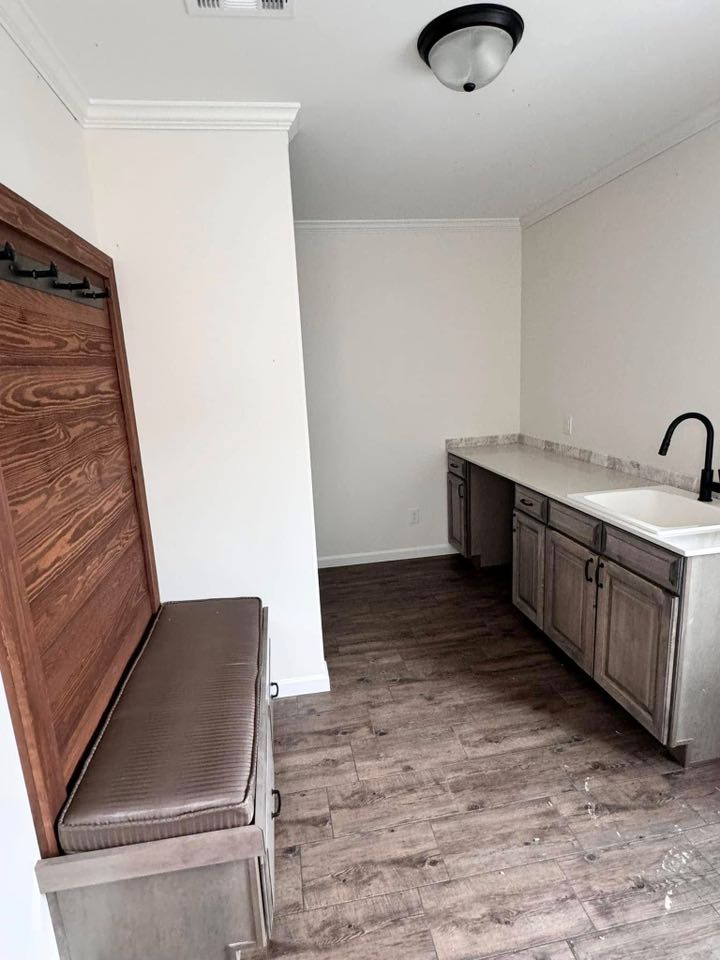 Small, modern laundry room with wood flooring, white walls, and dark cabinets. A cushioned bench with hooks sits on the left, and a sink with a black faucet is to the right.