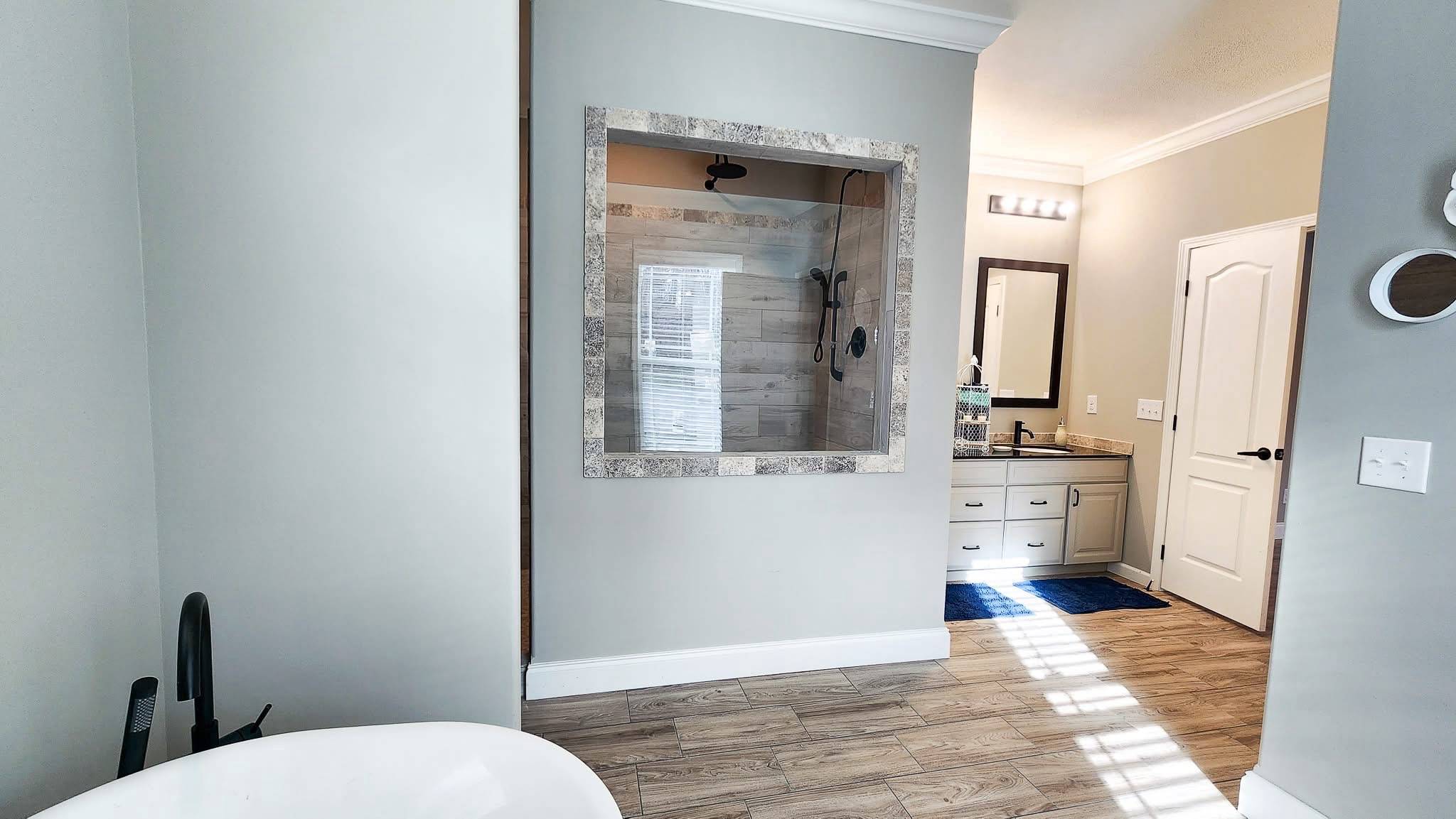 Modern bathroom with a freestanding tub, wood-look tile floor, and a large glass-enclosed shower. A vanity with a mirror is on the right. Bright and serene.