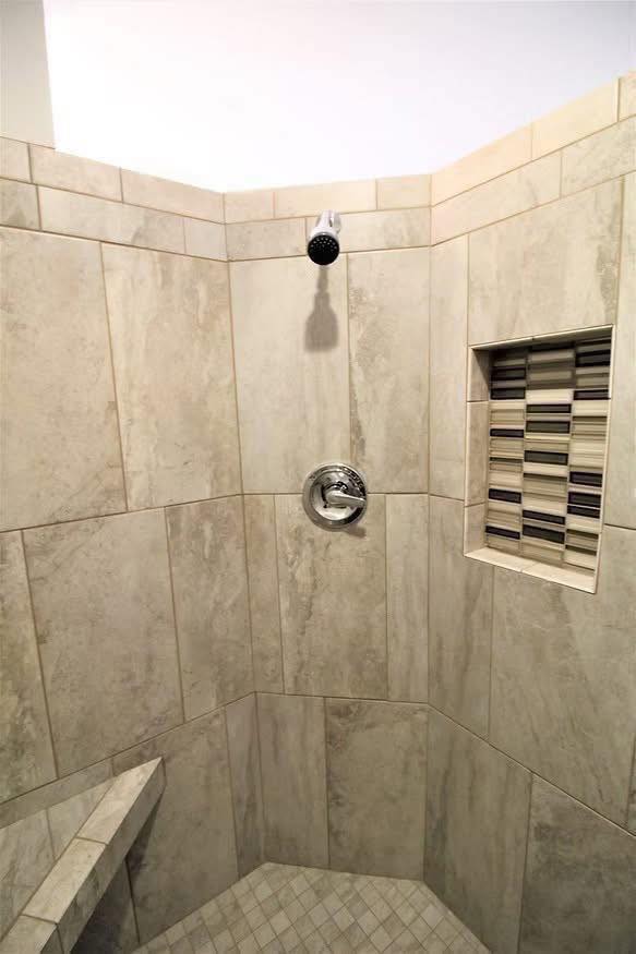 Tile shower with beige and cream ceramic tiles. Features a metal showerhead and faucet, a mosaic-tiled niche for storage, and a small corner bench. Clean, modern aesthetic.