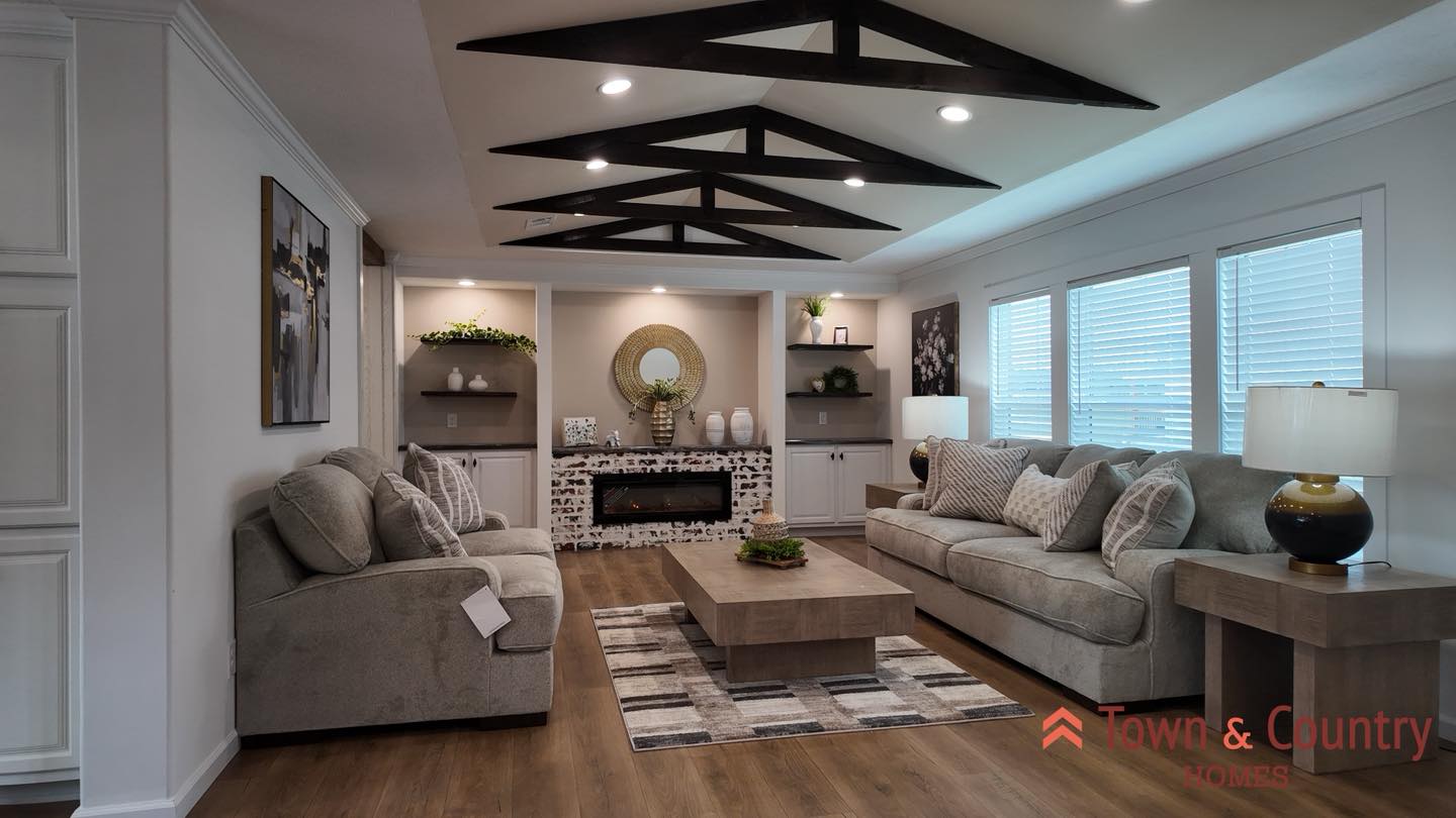Cozy living room with rustic beams, gray sofas, a central wooden coffee table, and decorated fireplace. Modern decor with natural light from large windows.