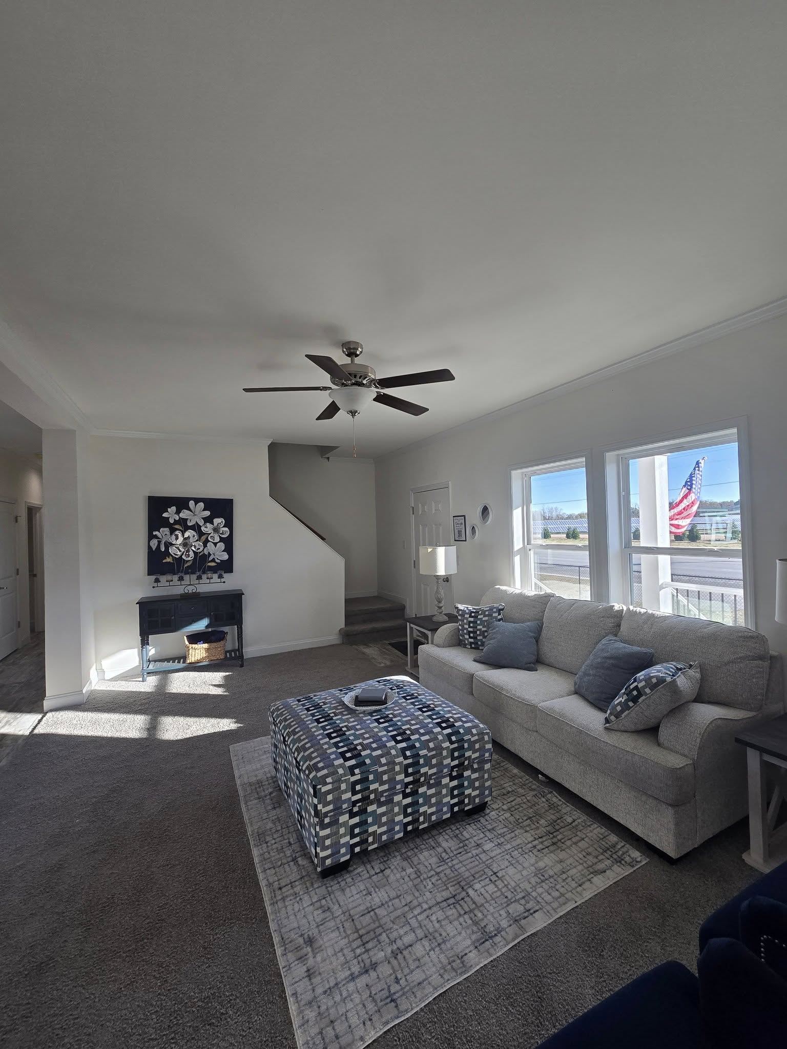 A cozy, sunlit living room with a gray sofa, blue-patterned ottoman, and elegant floral artwork. Large windows reveal a distant American flag.