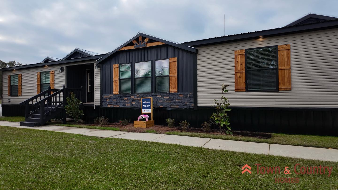 A modern single-story home with dark and light siding, wooden shutters, and a small front garden. The entrance features a black stairway. "Town & Country" logo is visible.