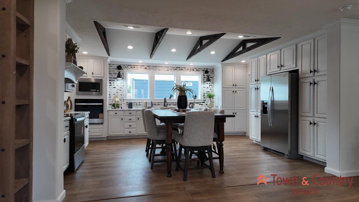 Spacious modern kitchen with wooden floors, white cabinets, and a central dining table. Large windows brighten the room, creating an inviting atmosphere.