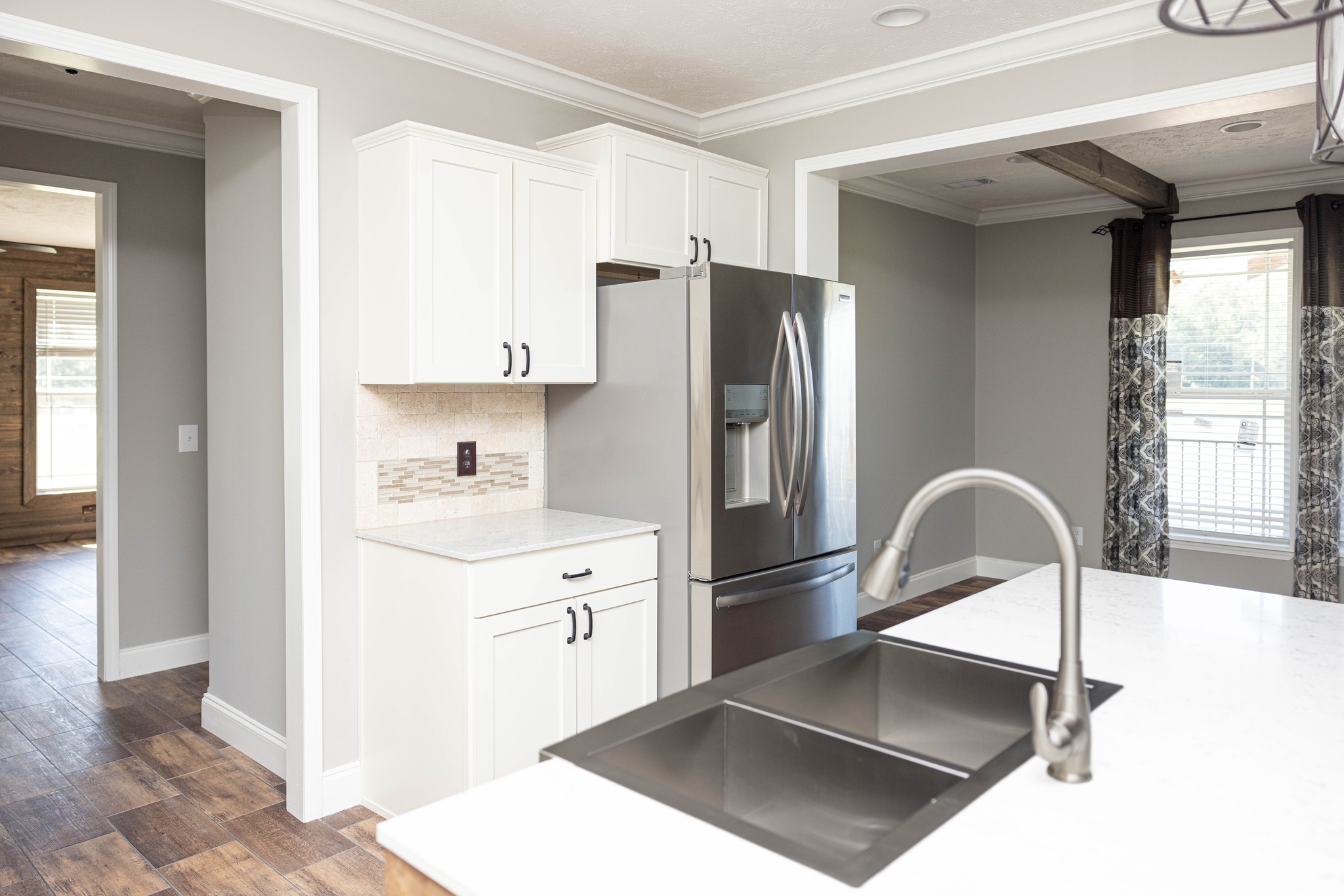 Modern kitchen with stainless steel fridge, white cabinets, and a sleek faucet over a large black sink. Sunlit, neutral tones, wooden floor in the background.