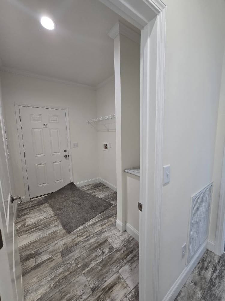 A narrow laundry room with gray wood-patterned tile flooring, a white door, and overhead shelving. Soft lighting creates a clean, minimalist tone.