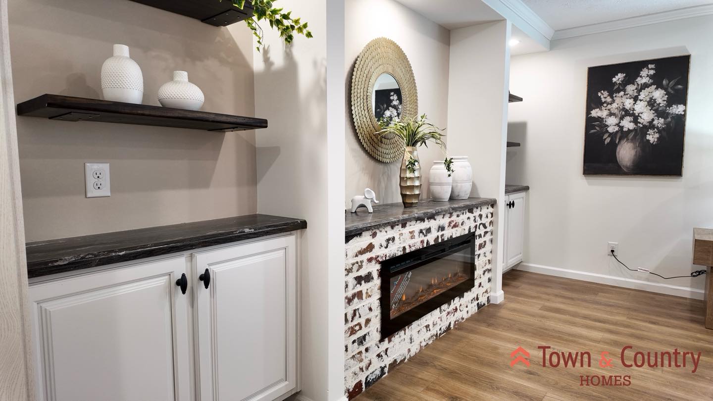 A cozy living room with a brick fireplace, round mirror, and potted plant. Shelves display white vases. A floral painting hangs nearby on a light wall.