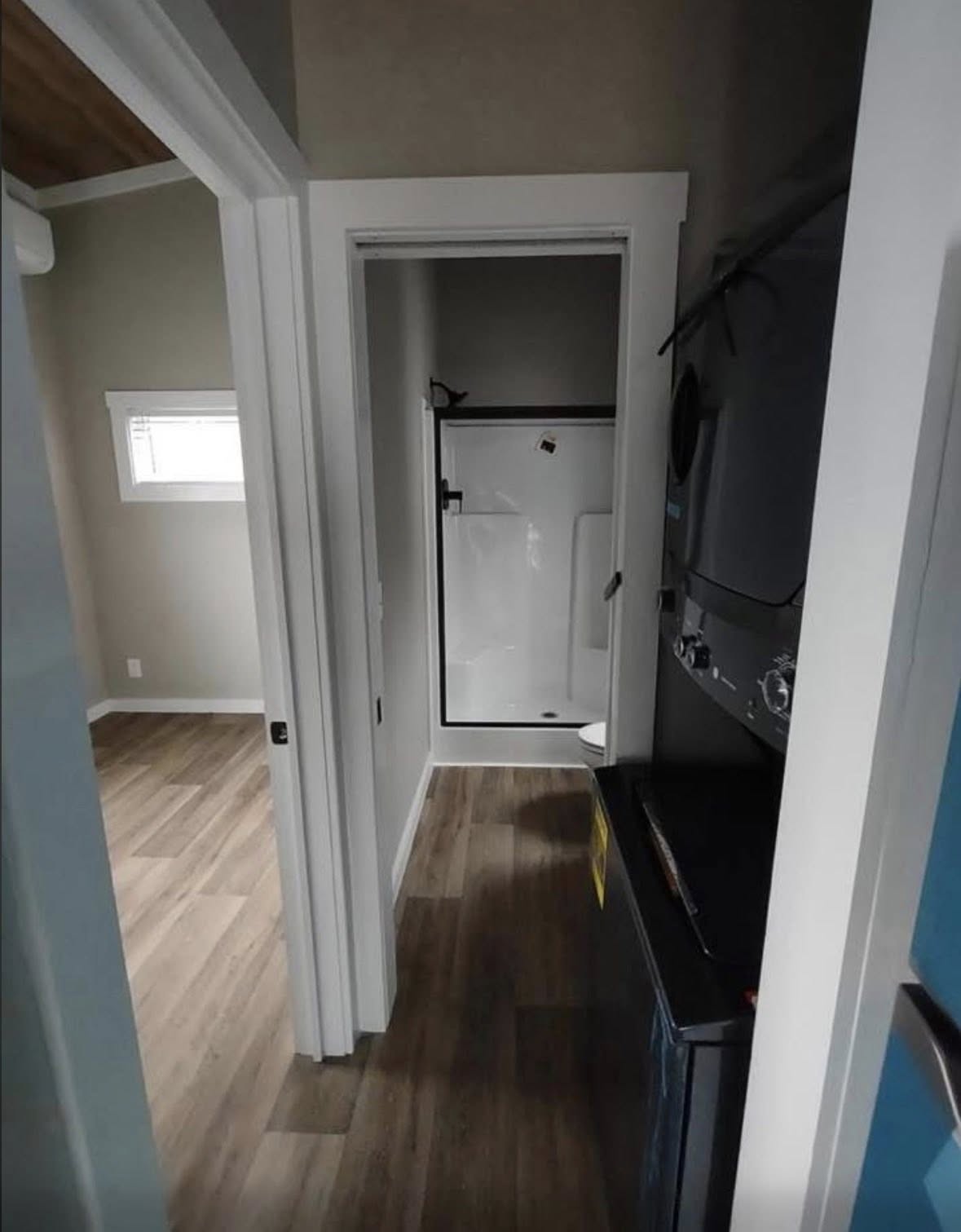 Narrow hallway with wood flooring, leading to a small bathroom with a glass-door shower. A stacked washer and dryer are on the right. Minimalist design.